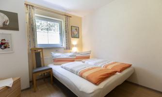 A cozy bedroom with a double bed and colorful pillows. The room has a window through which daylight streams, and is simply but cheerfully furnished.