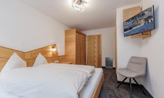 A modern bedroom with a double bed and wooden wardrobes. There is a television on the wall and a comfortable chair in the corner.