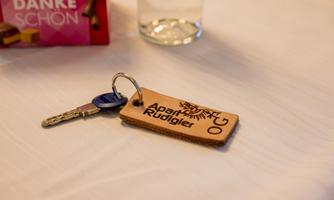 A keychain with a leather key fob displaying the words "Apart Rüdigler". In the background, there is a glass and a box of sweets.
