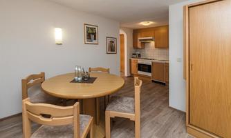 A cozy dining area with a round wooden table and four chairs. In the background, there is a modern kitchen.