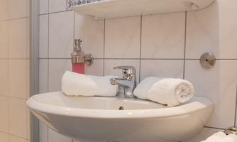 A modern bathroom with a white sink, two towels, and a large mirror. The walls are tiled in light colors and there is a shower in the background.