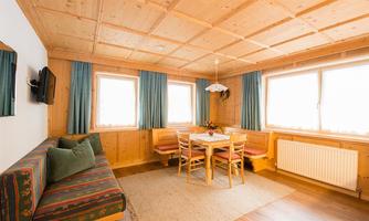 A cozy living space with wood paneling and large windows. In the center, there is a table with chairs, surrounded by a couch and decorative elements.