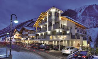 A modern hotel in the snow, surrounded by mountains. The facade is illuminated and there are parked cars in front of it.