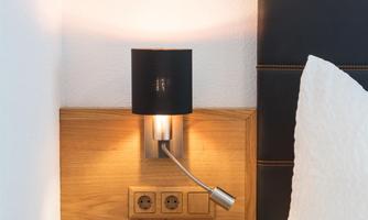 A cozy bedroom with a wooden nightstand. On the wall, there is a modern bedside lamp.