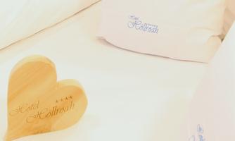 A wooden heart with the inscription "Hotel Hollbrath" lies on a bed. In the background, pillows with the hotel name are visible.