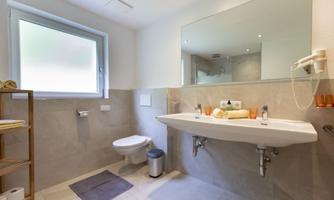 A modern bathroom with a bright floor and a large sink. There is a window, a toilet, and towels on an open shelf.
