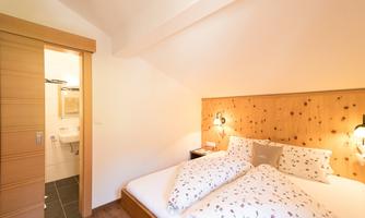 A cozy bedroom with a double bed and wood paneling. In the background, access to the bathroom is visible.