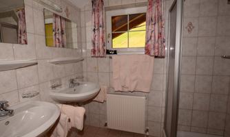 A bright bathroom with two washbasins, a shower area, and a window. The walls are tiled and decorated with pink towels and curtains.