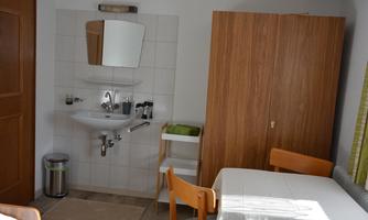 A simple room with a table, two chairs, and a sink. The walls are light, and there is a wooden cupboard as well as a trash can.