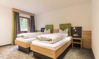 A modern bedroom with two single beds and light wooden furniture. Large windows with curtains create a pleasant lighting atmosphere.