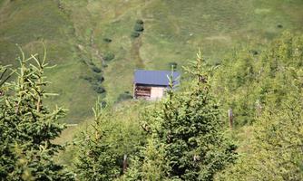 A small cabin stands in a green mountain landscape. Surrounded by dense trees and gentle hills.