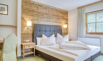 A modern bedroom with a cozy bed and rustic wood paneling. The room features a bright window front and pleasant lighting.