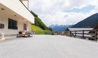 A bright terrace with chairs and a table offers a view of the surrounding mountains. The clouds over the peaks add a relaxed atmosphere to the scene.