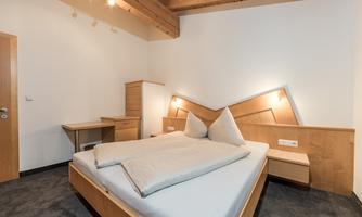 A modern bedroom with a double bed and wooden details. The walls are light, and there is a desk and bedside tables.