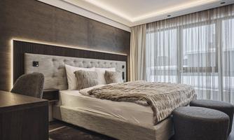 A stylish hotel room with a large bed and modern furniture. The room is bright and has large windows with curtains.