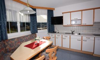 A cozy kitchen with a dining table and chairs. The walls are painted in light colors, and there is a television as well as windows with green curtains.