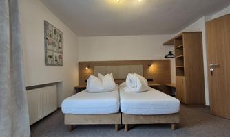 A cozy bedroom with two single beds and neutral colors. The decor is simple, with a shelf and a piece of artwork on the wall.
