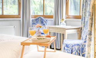 An elegant room with light curtains and blue furniture. On a table, there are two glasses of orange juice and a breakfast plate.