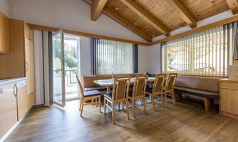 A bright dining room with a large wooden table and chairs. The windows are open and offer a view of nature outside.