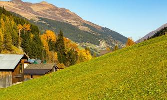 A picturesque mountain landscape with green meadows and colorful autumn foliage. In the background, the mountains and small houses can be seen.