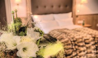 A cozy bedroom with a large bed and soft lighting. In the foreground, a beautiful bouquet is visible.