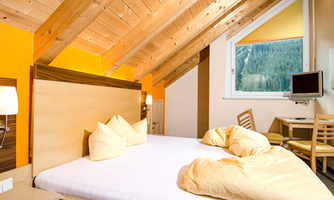 A cozy bedroom with a large bed and wooden ceiling. The room has windows overlooking nature and a small workspace.