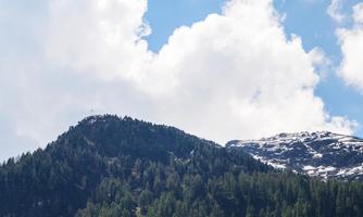 A beautiful mountain with some snow-covered areas and a green forest landscape. The sky is partly cloudy and radiates brightness.