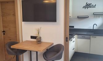 A modern dining room with a small table and two chairs. In the background, a kitchenette and a television are visible.