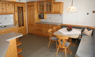 A cozy kitchen with wooden cabinets and a dining area. On the table, there is a floral tablecloth and a pleasant atmosphere.