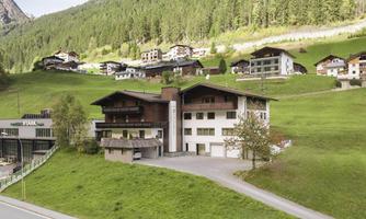 A modern building in a green environment with several other houses in the background. The landscape is hilly and surrounded by trees.