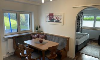 A cozy dining area with a wooden table and a cushioned seating area. In the background, there is a bedroom with a large bed.