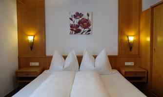 A cozy bedroom with a double bed and decorative pillows. A painting of blooming flowers hangs on the wall, and the walls are made of light wood.