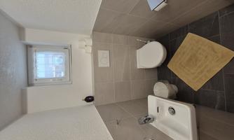A small, modern bathroom with a sink and a toilet. The room has bright tiles and a window for daylight.