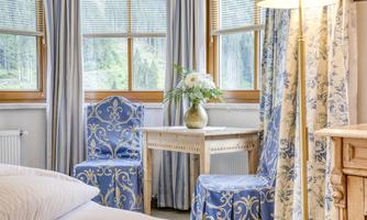 A cozy room with blue chairs and a small table.  
On the table stands a vase with flowers and large windows let in plenty of light.
