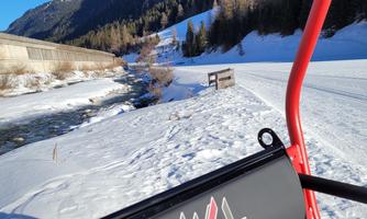 An idyllic winter view of snow-covered mountains and a clear river. In the foreground, a chairlift with the inscription "Ischgl" is visible.