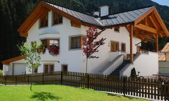 A modern house with a wooden roof and white walls is situated in a green landscape. The garden is surrounded by a fence, and there are some trees and plants.