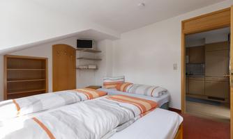 A simple bedroom with two single beds and orange bedspreads. In the background, an open kitchenette can be seen.