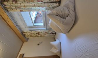 A cozy bedroom with a large bed and decorative cushions. The windows are adorned with floral curtains and provide a view of natural light.