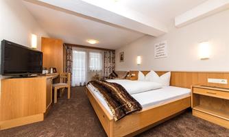 A cozy hotel room with a large bed, a television, and a desk. The walls are light, and there are windows with curtains for daylight.
