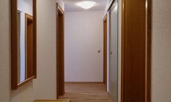 A narrow hallway with bright walls and a warm wooden floor. On the left side, there is a mirror and a wooden bench.