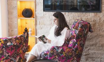 A person in a white bathrobe sits relaxed in a colorful armchair and reads a book. In the background, a television and a decorative wall with shelves can be seen.
