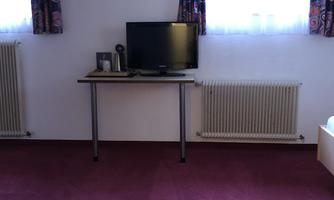 A simple room with a television on a table and two windows with curtains. The floor is covered with red carpet.