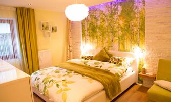A cozy bedroom with a large bed and colorful pillows. The wall is decorated with a tree motif and the room has warm, inviting lighting.
