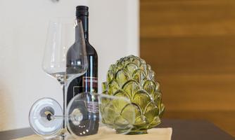 A bottle of red wine stands next to two wine glasses and a decorative green bowl in the shape of an artichoke. The composition creates an elegant and inviting atmosphere.