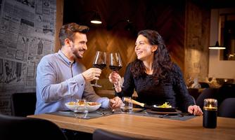 A couple sits in a stylish restaurant and looks at each other with smiles. They clink their wine glasses and enjoy their dinner.