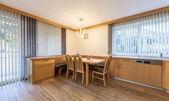 A modern kitchen with wood decor and a dining area. Large windows and blinds provide plenty of light in the room.