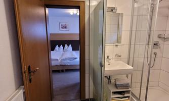 A modern bathroom with a shower and a sink. In the background, a bedroom with a bed and white pillows is visible.