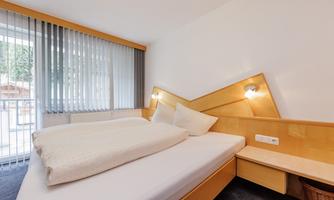 A modern bedroom with a large bed and a bright decor. The windows are covered with blinds and let in daylight.