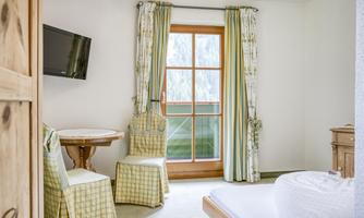 A cozy room with a window that lets in plenty of daylight. There is a table, two chairs, and a television on the wall.