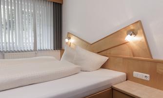 A modern bedroom with a wooden bed and two pillows. The walls are light and the windows have vertical blinds.